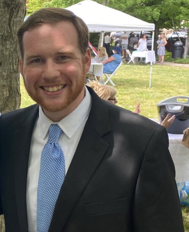 Zacherie Kinslow is a white man in a black suit with a white shirt and light blue tie. He is standing outside, in front of a picnic.
