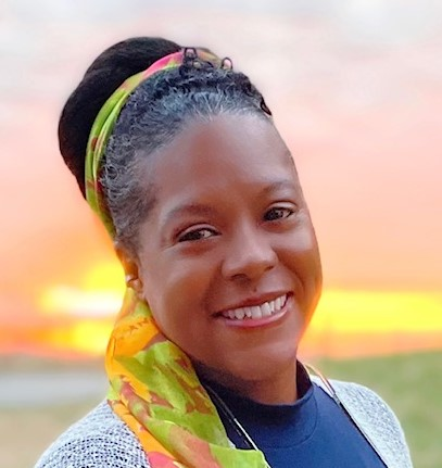 Headshot of Jemma Perran, and African-American woman in a navy blue shirt and white sweater. Her hair is tied with a multicolored scarf. She is standing in front of a pink and yellow sunset.